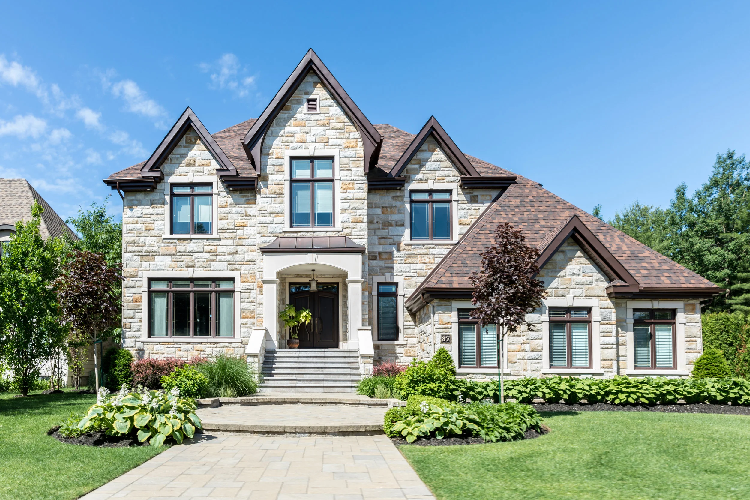 A large stone house with triangular roof peaks, white-framed windows, and a manicured lawn. The sky is clear and blue, conveying a serene atmosphere.