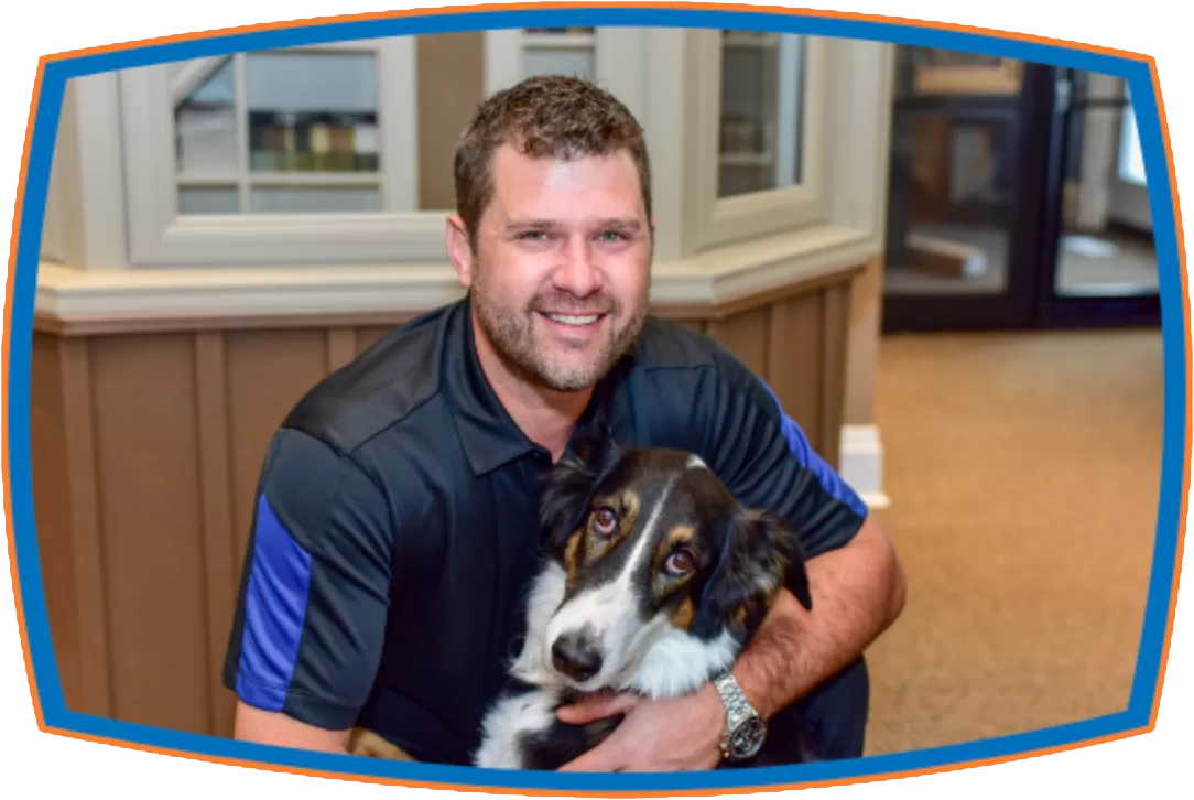 Chris in a black polo shirt smiling warmly while hugging a black and white dog indoors. The setting is cozy, conveying a sense of friendship and comfort.
