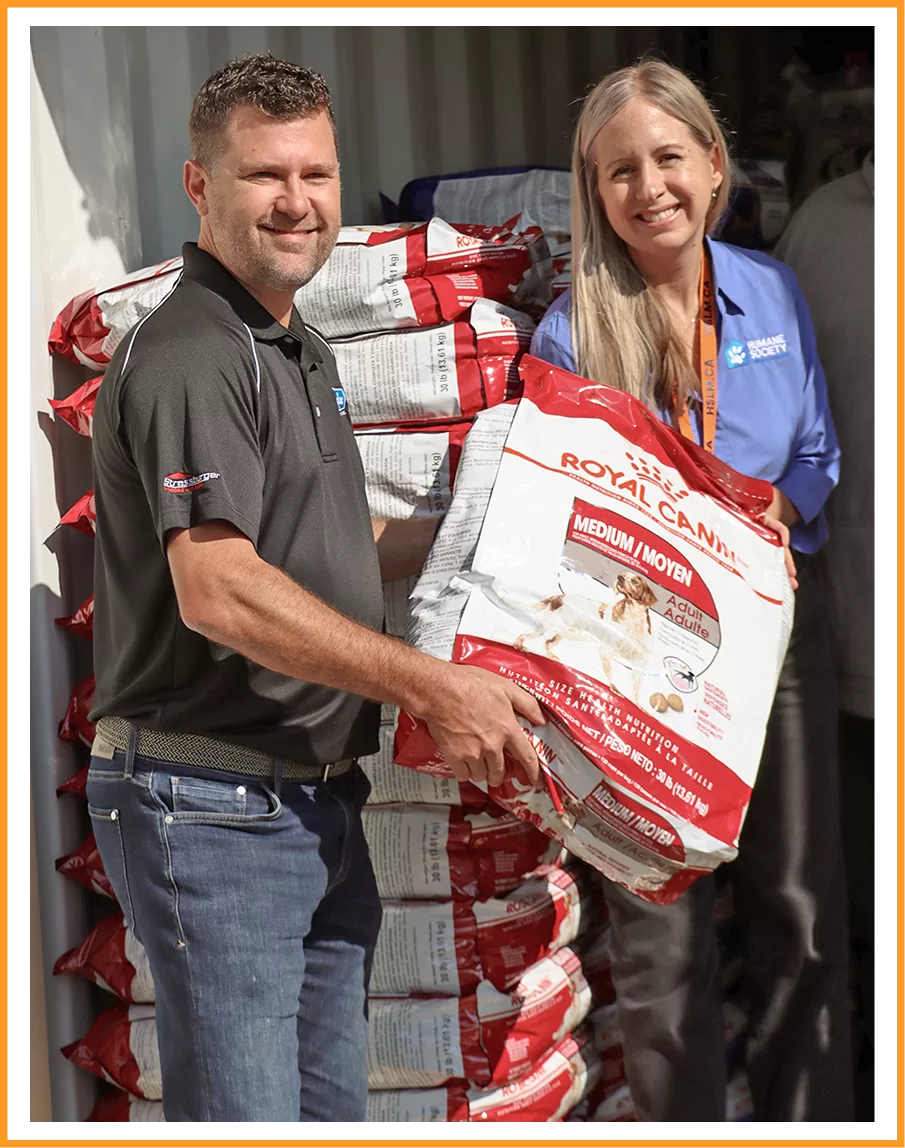 Two people smiling, holding a large bag of Royal Canin dog food. More dog food bags are stacked in the background. Bright, cheerful setting.