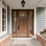 A welcoming home entrance with a rich wooden front door and glass sidelights, white siding, brick planters, colourful flowers, and a stone porch.