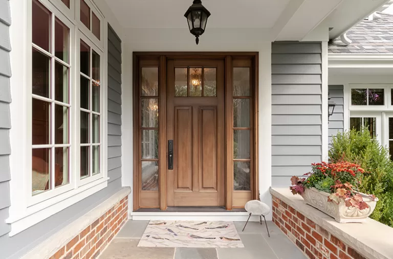 A welcoming home entrance with a rich wooden front door and glass sidelights, white siding, brick planters, colourful flowers, and a stone porch.